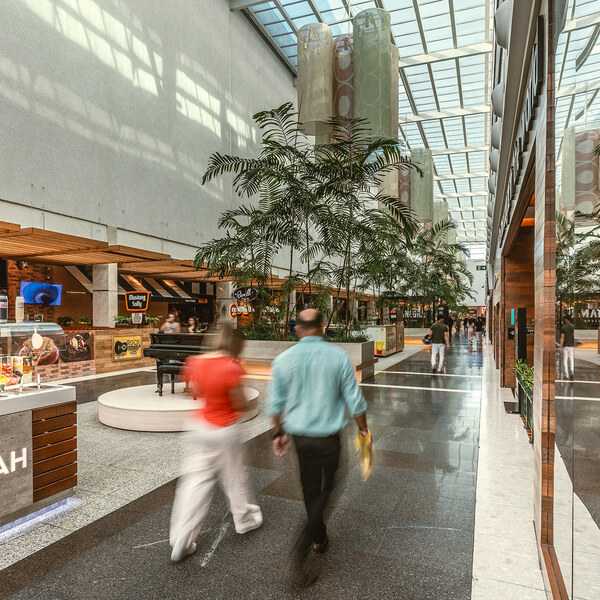 People walking in a bright indoor shopping mall corridor with plants, glass ceiling, and store fronts.